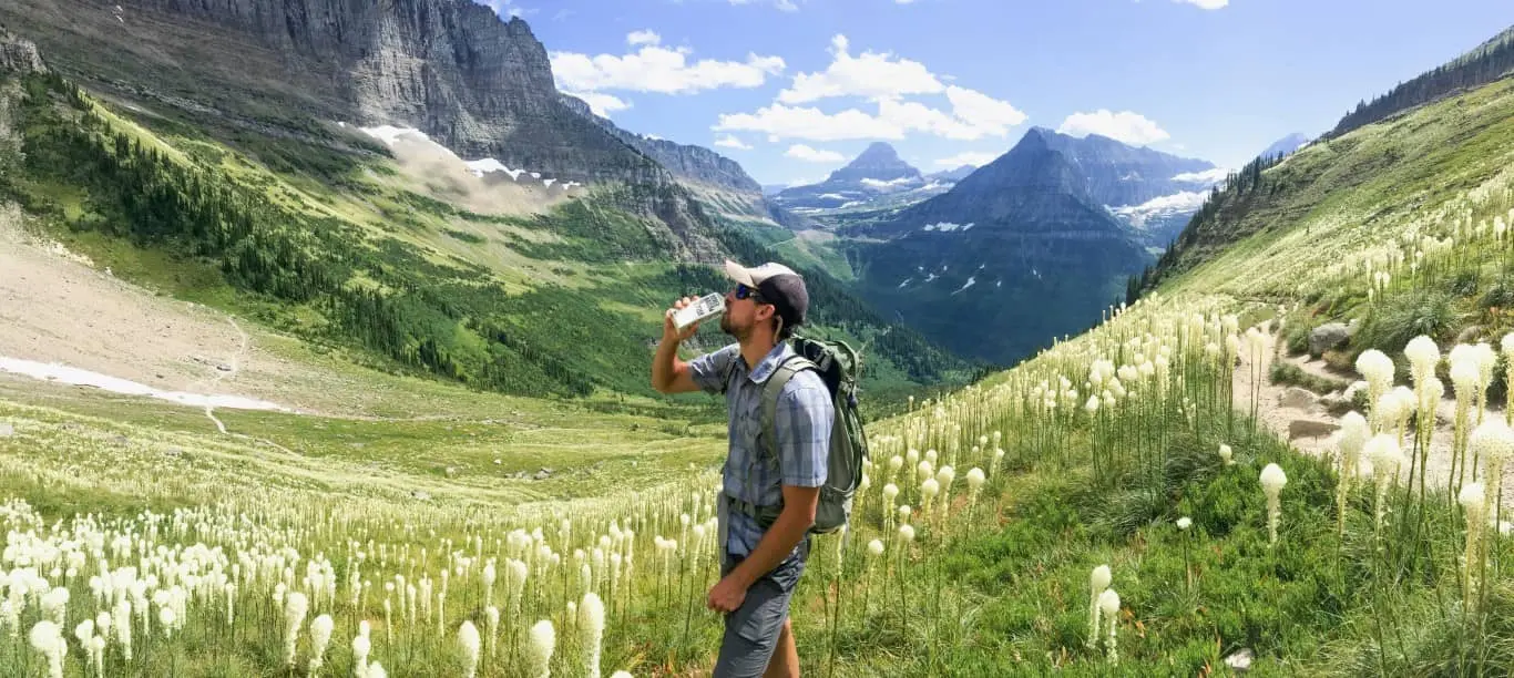 person drinking from his bottle in a grassy hillside with mountains in the background