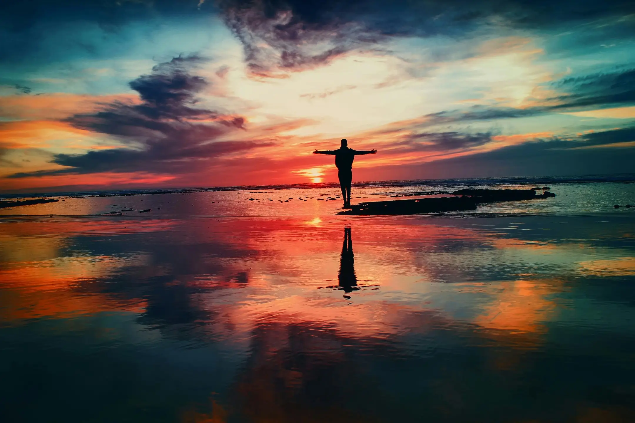 person standing on a shore in the sunset with a red light in the sky and reflecting on the water surface